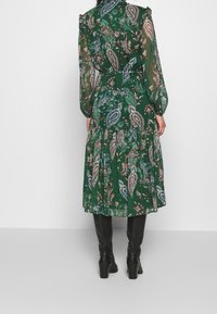 Mujer vestida con un vestido de manga larga de patrón paisley verde y botas de tacón negras hasta la rodilla, de pie frente a un fondo blanco liso.