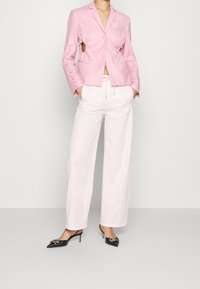 Rosa blazer met uitsnijdingen, gecombineerd met losse witte broeken en zwarte puntige hakken met een accent. Glad textuur overal.