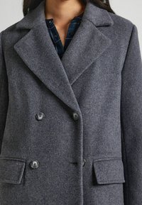 Abrigo de mezcla de lana gris, de doble botonadura, con solapas anchas, dos botones y dos bolsillos frontales. Tela texturizada y diseño ajustado.