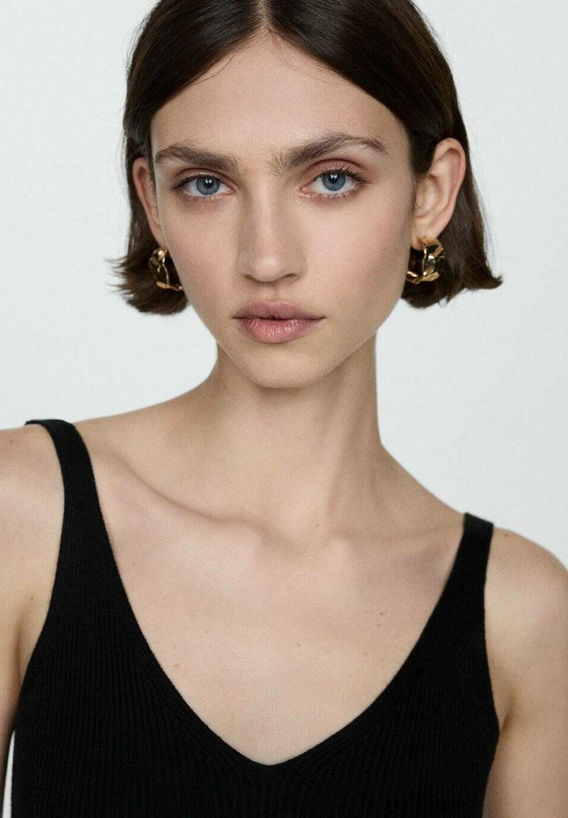 Młoda kobieta z krótkimi brązowymi włosami, niebieskimi oczami, nosząca złote kolczyki w kształcie kół i czarny top bez rękawów, spoglądająca na wprost na jednolitym tle.