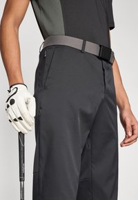 Šedé golfové kalhoty s texturovaným pasem a černým zapínáním. Bílá rukavice drží černou golfovou hůl, která ukazuje elegantní, moderní design.