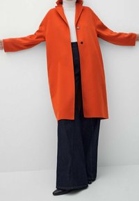 Un lungo cappotto arancione brillante con colletto, chiusura a un bottone e maniche larghe, abbinato a pantaloni di jeans scuri a zampa d'elefante e scarpe nere.