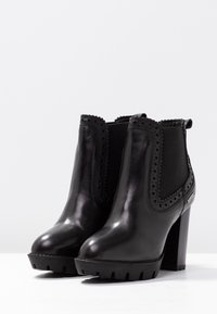 Botas de tobillo de cuero negro con un tacón alto y robusto, decoración de perforaciones, paneles elásticos en los laterales y una suela texturizada.
