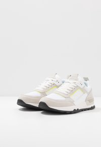 Baskets blanches en mélange de suède et de mesh, avec des accents gris et une ligne jaune néon. Semelle en caoutchouc avec un dessous noir texturé.