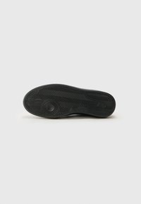 Black sneaker sole with a textured rubber finish, featuring a herringbone pattern and circular logo. Smooth edges and reinforced toe area.