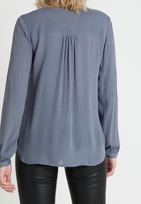 Blouse à manches longues gris clair avec une finition texturée, dotée d'un détail de pli le long du dos et de poignets à boutons. Associée à un pantalon noir.