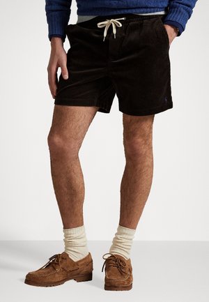 Man draagt zwarte corduroy short met wit trekkoord, crèmekleurige sokken, bruine suède veterschoenen en een blauwe trui met één hand in de zak.