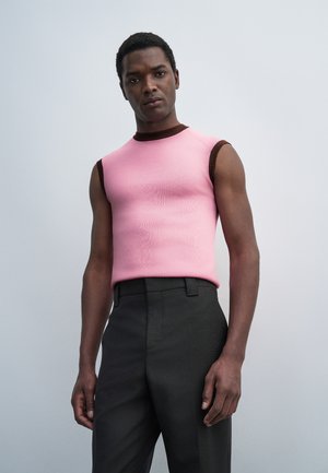 Homme portant un haut rose sans manches avec une bordure sombre et un pantalon noir, debout devant un fond gris clair uni.