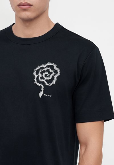 Jeune homme portant un t-shirt noir avec un motif de rose blanche effilochée et le texte "NN, 07" sur la poitrine gauche.
