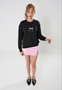 Svart genser med et rosa sløyfedesign på brystet, kombinert med en lys rosa mini-skjørt med side-slitt, og svarte spisse leiligheter.