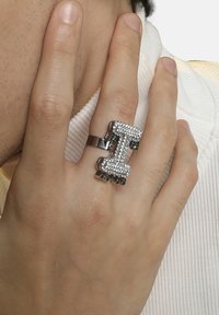 Sølvring med et dristigt, rektangulært design udsmykket med klare krystaller og grønne accenter, båret på en finger mod en tekstureret baggrund.