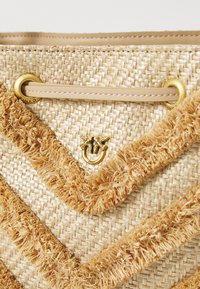Geweven stro tas in beige, met een franjes ontwerp en goudkleurige hardware. Details zijn onder andere een bij-embleem en decoratieve bovenstiksels.