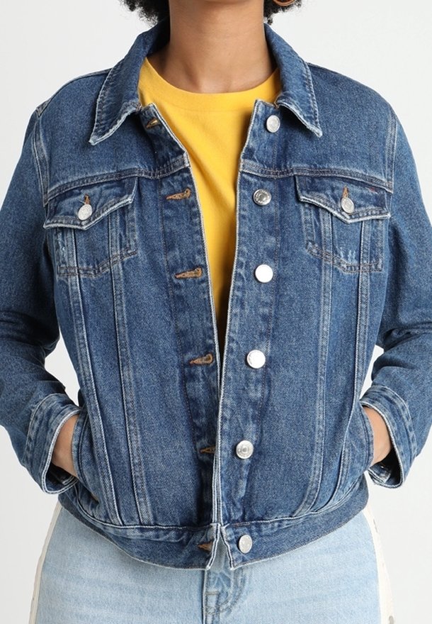 Veste en denim avec une coupe classique, couleur bleu clair, devant à boutons et deux poches poitrine ; portée sur une chemise jaune. Tissu structuré et texturé.