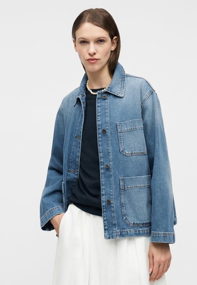 Γυναίκα που φοράει ανοιχτό μπλε τζιν μπουφάν με μεγάλες τσέπες, μαύρο τοπ, λευκή φούστα, κολιέ με πέρλες, στέκεται μπροστά σε απλό φωτεινό φόντο.