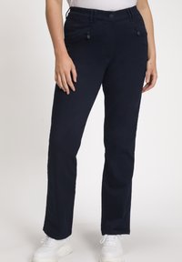 Pantalon droit marine avec fermeture éclair à l'avant et poches à boutons, porté avec des baskets blanches et un haut blanc rentré.