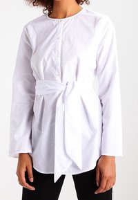 White button-up shirt with long sleeves and a tied waist. Features a round neckline and smooth, lightweight fabric.