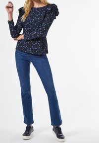 Schwarzes, floral bedrucktes Langarmoberteil mit rüschenbesetzten Schulterakzenten, kombiniert mit dunkelblauen, geraden Jeans und schwarzen Sneakers mit weißen Sohlen.