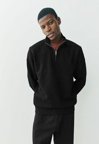 Sort zip-up sweatshirt med teksturerede træmønstre, lange ærmer og ribbet bund. Kombination med sorte bukser for et sammenhængende look.