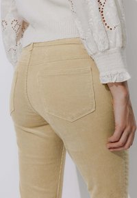 Pantalon en velours côtelé beige clair avec une texture côtelée, doté de deux poches arrière et d'une coupe ajustée, associé à un haut blanc brodé.