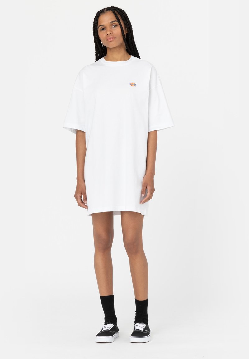 Biała, oversize'owa sukienka t-shirtowa wykonana z bawełny, z krótkimi rękawami i małym haftowanym logo na piersi. Połączona z czarnymi skarpetkami i butami.