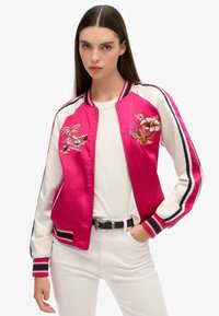 Veste bomber en satin de couleur rose vif avec des manches blanches, ornée de motifs floraux brodés et de accents sportifs rayés sur les poignets et le col.