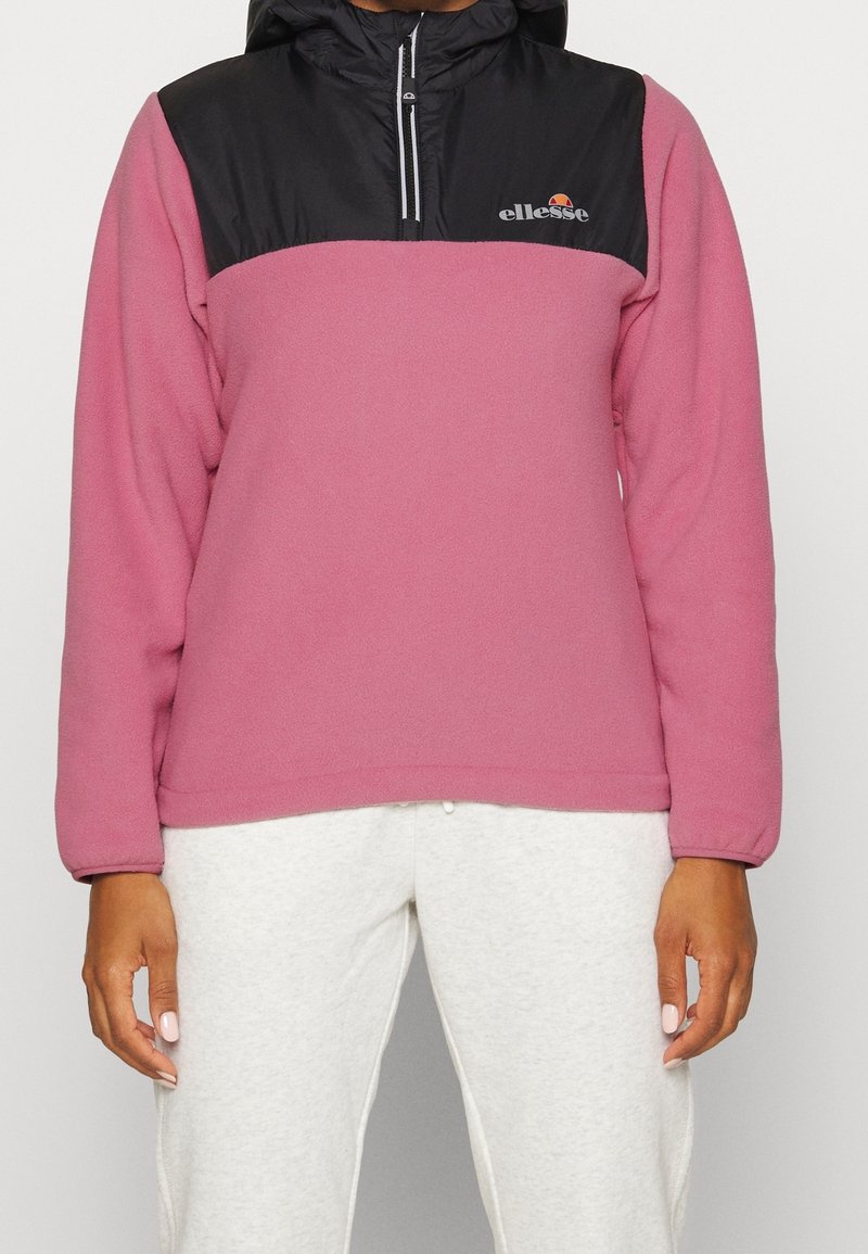 Pullover in pile rosa con una sezione superiore nera con cappuccio, zip frontale a un quarto e logo ricamato sul petto. Tessuto morbido in tutto il capo.