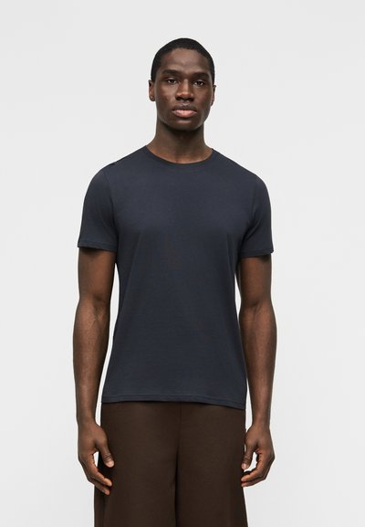 Jeune homme debout, face à l'avant, portant un t-shirt à manches courtes bleu marine uni et un pantalon marron, sur un fond clair.