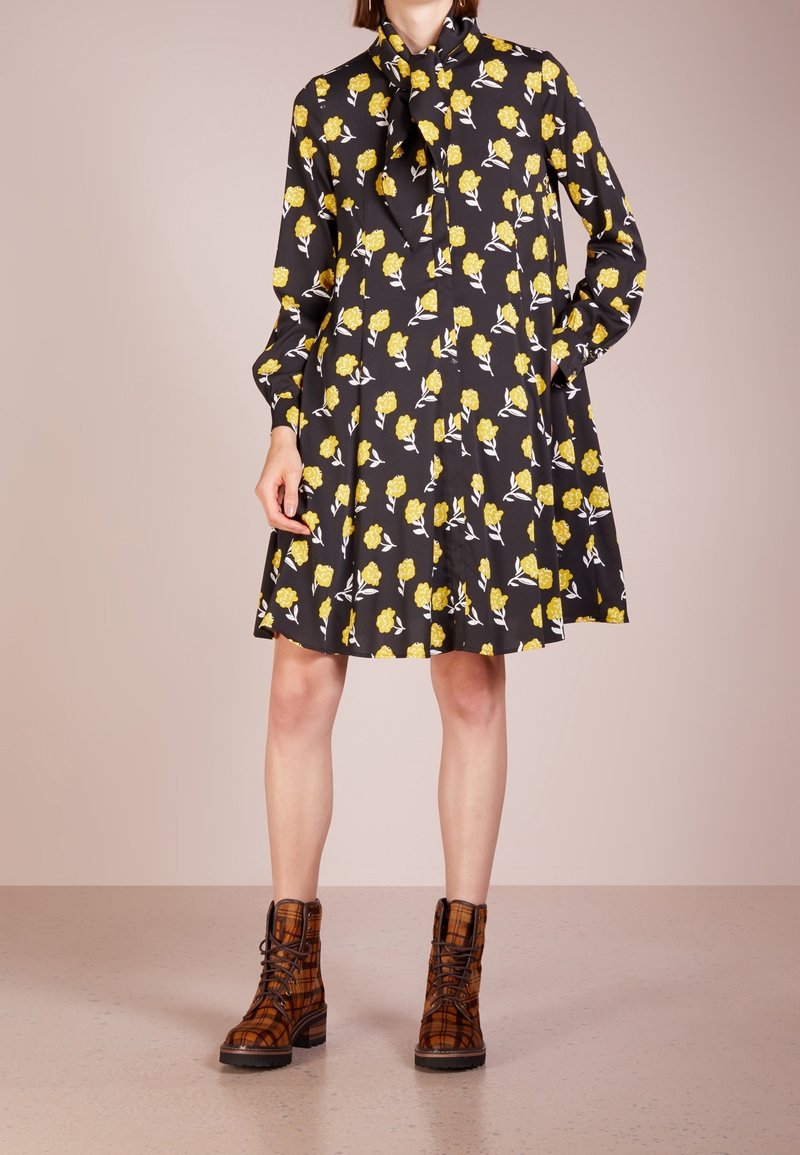 Svart klänning med gult blommönster, långa ärmar och knytkrage. Matchas med bruna rutiga ankelskålar med snörning och kraftiga sulor.