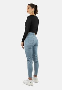 Schwarzes langärmliges Crop-Top, kombiniert mit hellblauen High-Waist-Jeans. Modell trägt weiße Sneakers, hat einen Pferdeschwanz und goldene Ohrringe.