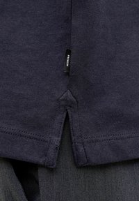 Dunkel marineblaue Stoffkante mit einem kleinen schwarzen Etikett mit der Aufschrift „PREMIUM“ in der Nähe eines seitlichen Schlitzes, überlagert von dunkelgrauem, strukturiertem Stoff darunter.