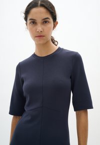 Námořnické modré šaty s krátkými rukávy, přiléhavý střih, kulatý výstřih a decentní švy na látce. Hladká textura, minimalistický styl.