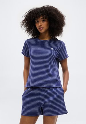 Kobieta o kręconych włosach, ubrana w granatowy, pasiasty krótki top z krótkim rękawem i dopasowane szorty, z rękami w kieszeniach, neutralne tło.