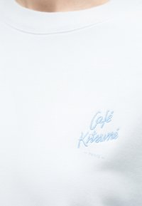 Κοντινό σε λευκό ύφασμα με κεντημένο με γαλάζιο κείμενο "Café Kitsuné - Paris" στην περιοχή του στήθους.