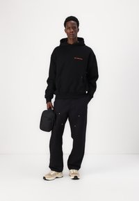Schwarzer Hoodie mit orangefarbigem Text "Verschwende keine Kultur", schwarze Hose mit sichtbaren Nähten und weißen Akzenten, beige und schwarze Sneakers, hält eine kleine schwarze Tasche.