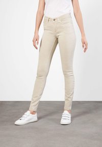 Jeans skinny beige con un diseño clásico de cinco bolsillos, cierre frontal con botón y detalles de costura sutiles, combinados con zapatillas blancas.