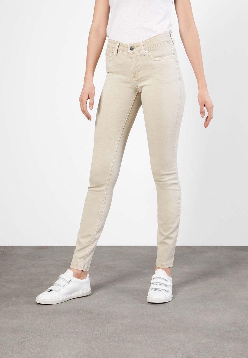 Jeans skinny beige con un diseño clásico de cinco bolsillos, cierre frontal con botón y detalles de costura sutiles, combinados con zapatillas blancas.