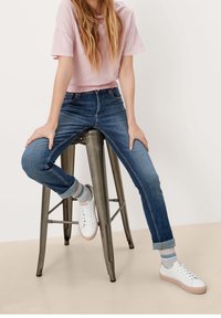 Ljusrosa croppad t-shirt, mörkblå tajta jeans, grå och rosa randiga strumpor, vita sneakers med rosa sulor, sittande på en metallpall.