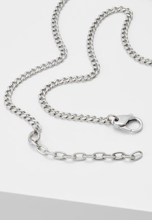 Collier chaîne en métal argenté avec maillons curb, fermoir mousqueton et chaînette d’extension courte posé sur une surface blanche.