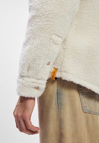 Lodden hvit lammeskinnsjakke med trykknapper og en gul etikett, brukt over lys beige denimbukser. Myk tekstur og uformelt design.