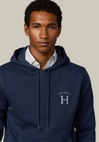 Sudadera con capucha de color azul marino, fabricada en un tejido suave, con un bolsillo frontal, que presenta un logo plateado y cordones con puntas metálicas.