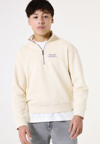 Sudadera de forro polar color crema con media cremallera y texto azul en el pecho, puños acanalados y un dobladillo estructurado sobre una camiseta blanca.