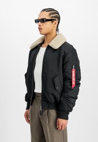 Cazadora bomber negra con un cuello de piel sintética beige, cierre de cremallera, bolsillos laterales y detalle de etiqueta roja en la manga izquierda. Llevada sobre una camisa blanca.