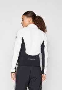 Valge vale karusnahast jakk mustade kangapaneelidega, kõrge kaelusega ja logode detailiga seljal; omab tekstuurset pinda ja sportlikku disaini.