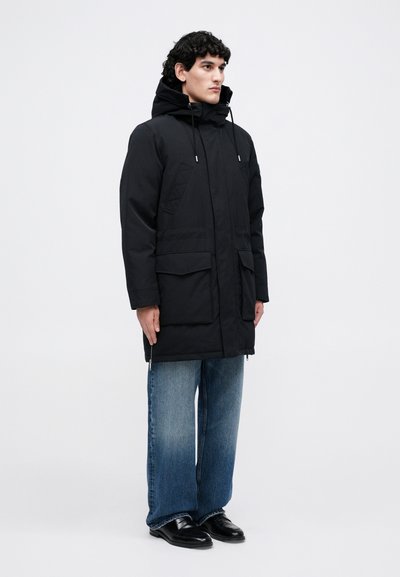 Juoda parka striukė su kapišonu, dviem priekinėmis kišenėmis ir užtrauktuku. Dėvima ant laisvo kirpimo mėlynų džinsų ir juodų batų. Lygi tekstūra.