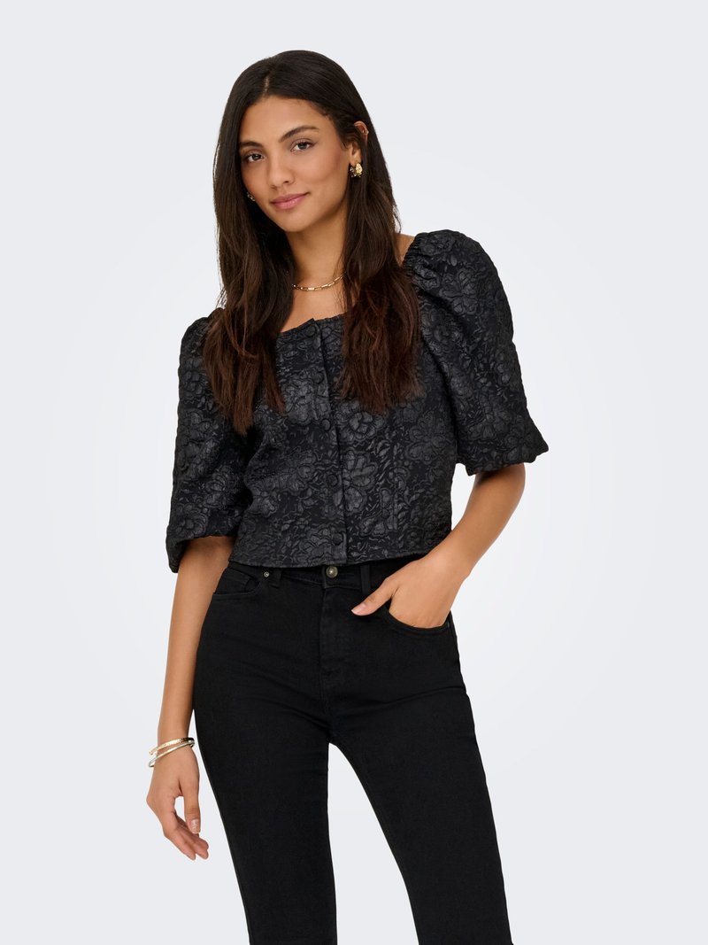 Femme aux longs cheveux foncés portant un chemisier noir à manches bouffantes texturé floral et un pantalon noir taille haute, posant avec une main dans la poche.