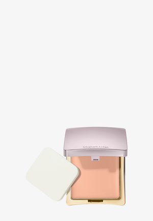 Fond de teint compact avec maquillage crème beige, couvercle argenté embossé du nom de la marque, et éponge applicatrice carrée blanche à côté.