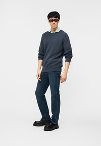 Νέος άνδρας με σκουρόχρωμα γυαλιά ηλίου, μπλε πλεκτό πουλόβερ πάνω από ανοιχτό πουκάμισο με γιακά, μπλε τζιν και μαύρα χοντρά παπούτσια, στέκεται σε λευκό φόντο.