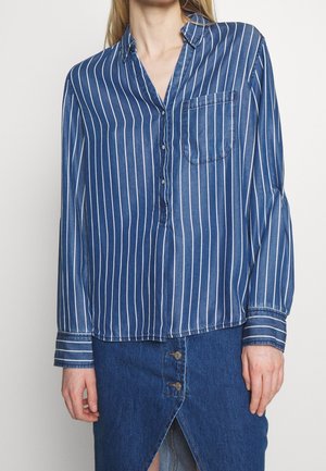 Vrouw draagt een blauw overhemd met verticale strepen, een borstzak en een blauwe spijkerbroekrok met een split aan de voorkant en knopen.