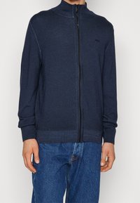 Man som bär en marinblå zip-up cardigan med ribbade ärmslut och nederkant, tillsammans med löst sittande blå denimjeans.