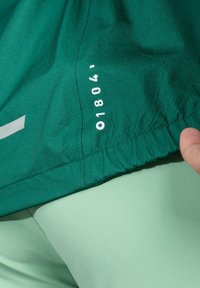 Zielona fakturowana tkanina z białymi cyframi i logotypem. Posiada elastyczny ściągacz i jest połączona z jasną miętową tkaniną pod spodem.